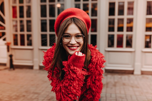 Jovem bronzeada com batom vermelho em óculos chapéu vermelho e casaco de lã sorri enquanto caminhava ao longo do prédio com janelas de chão