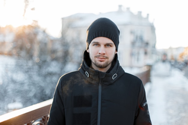 Jovem brutal com uma barba em um chapéu preto elegante com uma jaqueta de inverno nas árvores de inverno na cidade. Cara atraente.