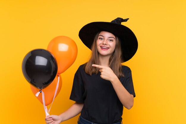 Jovem bruxa segurando balões de ar preto e laranja para festas de halloween, apontando para o lado para apresentar um produto