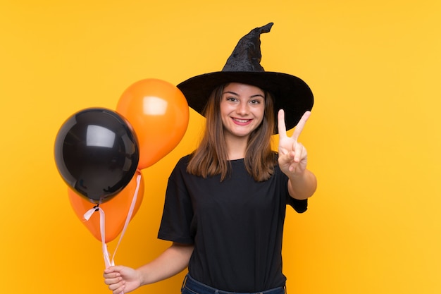 Jovem bruxa segurando balões de ar preto e laranja para festas de halloween, sorrindo e mostrando sinal de vitória