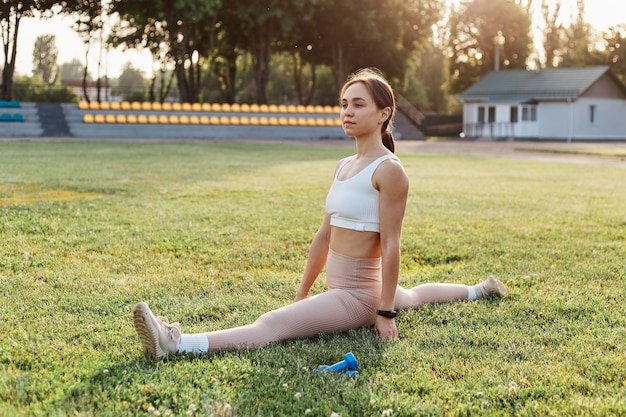 Jovem cabe mulher morena, fazendo barbante no estádio na grama verde. menina bonita, vestindo blusa branca e legging bege, esticando as pernas e o corpo do lado de fora no verão. treinamento de exercícios físicos.