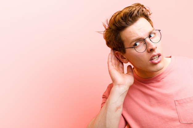 Jovem cabeça vermelha olhando sério e curioso, ouvindo, tentando ouvir uma conversa secreta ou fofoca, bisbilhotando a parede rosa