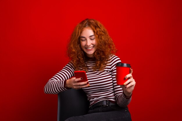 Jovem cabeça vermelha, sentado na cadeira e usando telefone celular enquanto segura uma xícara de café