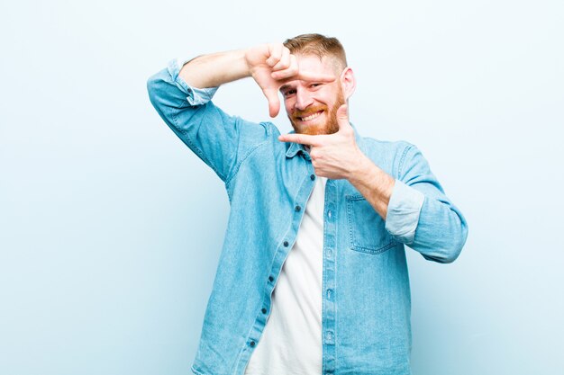 Foto jovem cabeça vermelha, sentindo-se feliz, amigável e positivo, sorrindo e fazendo um retrato ou moldura com as mãos no azul suave