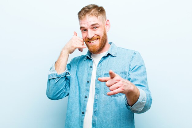 Jovem cabeça vermelha sorrindo alegremente e apontando para a câmera enquanto faz uma ligação, mais tarde, gesto, falando no telefone contra azul suave