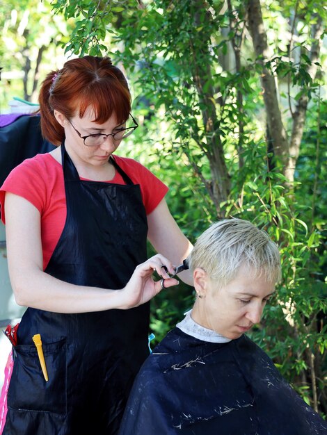 Jovem cabeleireira fazendo corte de cabelo curto com uma tesoura para uma mulher ao ar livre no jardim