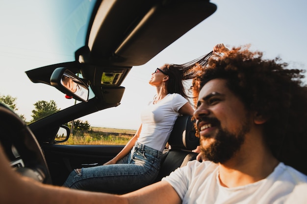 Foto jovem cacheado de cabelos escuros sentado ao volante de um cabriolet preto e uma linda mulher está sentada ao lado dele em um dia ensolarado. .