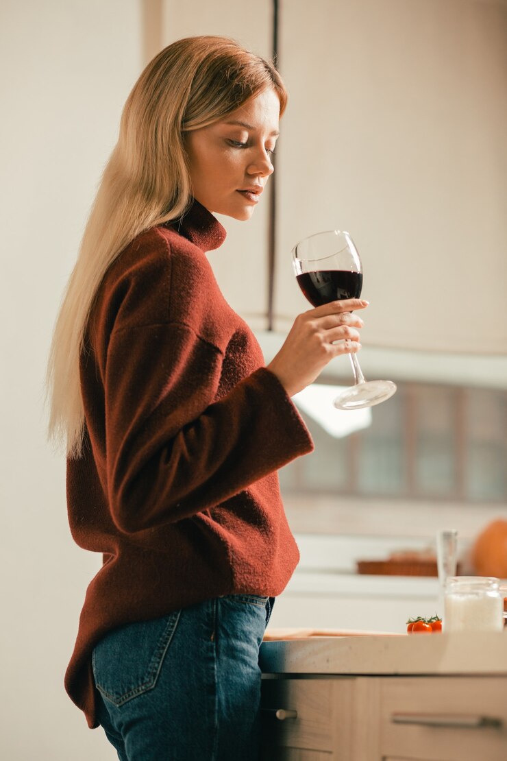 Jovem calma em pé na cozinha sozinha, olhando atentamente para o vinho tinto em seu copo