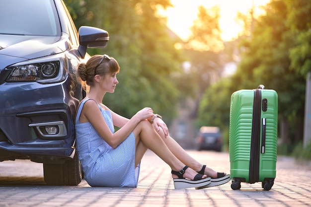 Jovem cansada com mala sentada perto de seu carro esperando por alguém Conceito de viagens e férias