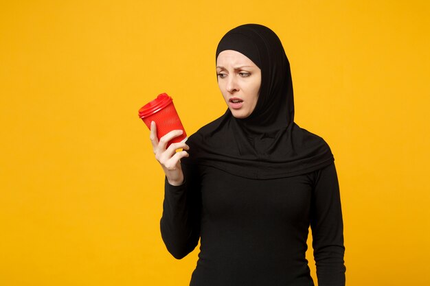 Jovem cansada triste chateada mulher muçulmana árabe em roupas pretas de hijab segurar o copo de papel de café isolado no retrato de parede amarela. Conceito de estilo de vida religioso de pessoas.