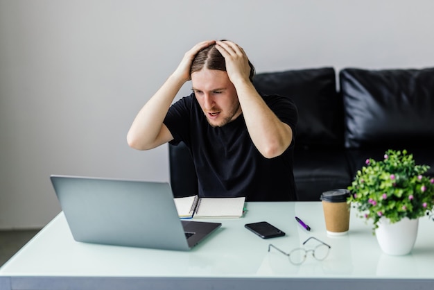 Jovem cansado sente dor, fadiga ocular, segurando os óculos, esfregando os olhos secos e irritados, cansado do trabalho no computador, homem estressado, sofre de dor de cabeça, problema de visão ruim, sente-se na mesa de casa usando o laptop