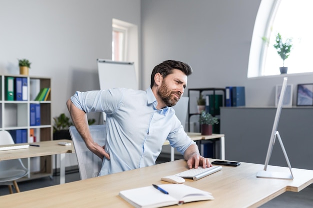 Jovem cansado trabalhador de escritório freelancer que trabalha no computador e se senta em uma mesa no escritório sente dor nas costas massageia suas costas relaxa