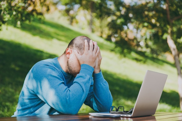 Jovem cansado triste homem empresário ou estudante em casual camisa azul, óculos, sentado à mesa com o celular no parque da cidade usando laptop, trabalhando ao ar livre, se preocupa com problemas. conceito de escritório móvel.