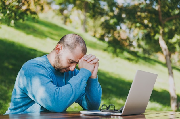 Jovem cansado triste homem empresário ou estudante em casual camisa azul, óculos, sentado à mesa com o celular no parque da cidade usando laptop, trabalhando ao ar livre, se preocupa com problemas. conceito de escritório móvel.