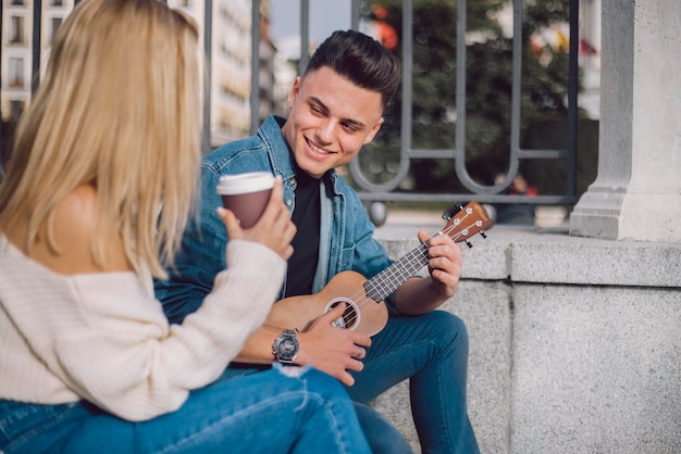 Foto jovem canta para seu parceiro e toca ukulele na rua ambos usam jeans e ela segura um café
