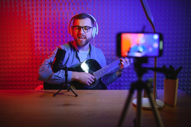 Jovem cantor caucasiano em fones de ouvido toca guitarra gravando novo single em smartphone em estúdio doméstico Artista milenar usa instrumento musical canta gravar videoclipe na câmera do celular