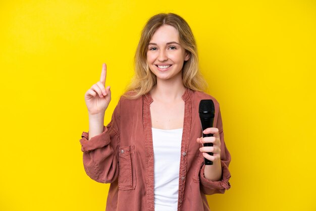 Jovem cantora caucasiana pegando um microfone isolado em fundo amarelo mostrando e levantando um dedo em sinal de melhor