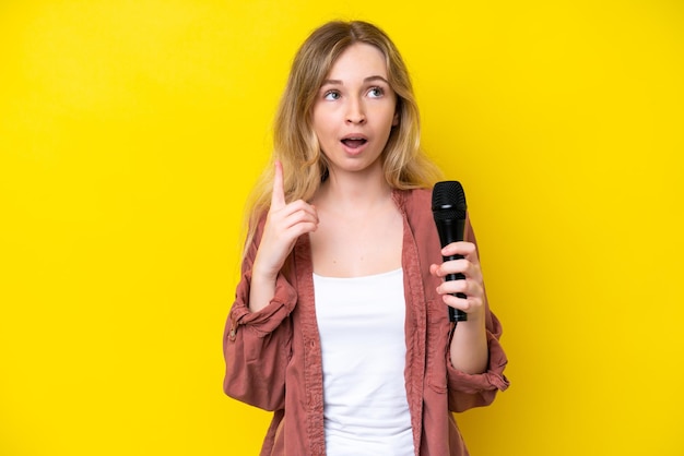 Jovem cantora caucasiana pegando um microfone isolado em fundo amarelo pensando uma ideia apontando o dedo para cima