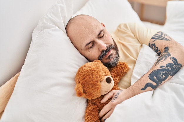 Jovem careca abraçando ursinho de pelúcia deitado na cama dormindo no quarto