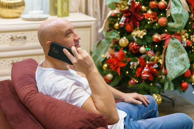 Jovem careca feliz senta-se em frente a uma árvore de natal decorada em seu apartamento e fala ao telefone ...