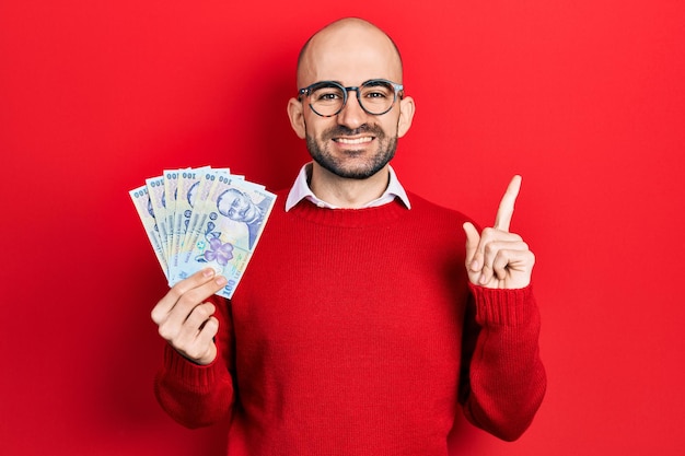 Jovem careca segurando 100 notas de leu romeno sorrindo feliz apontando com a mão e o dedo para o lado