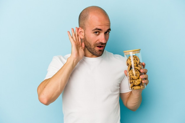 Jovem careca segurando biscoitos isolados sobre fundo azul, tentando ouvir uma fofoca.