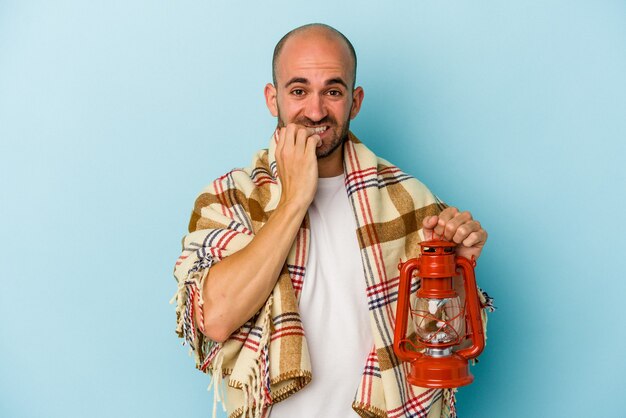 Jovem careca segurando lanterna vintage isolada em fundo azul, roendo as unhas, nervoso e muito ansioso.