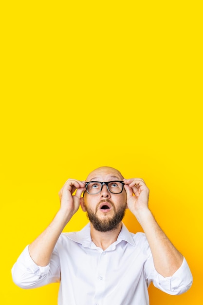 Jovem careca segurando óculos parece coceira em um fundo amarelo