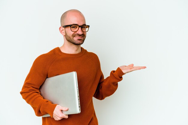 Jovem careca segurando um laptop isolado na parede branca, mostrando um espaço de cópia na palma da mão e segurando a outra mão na cintura.