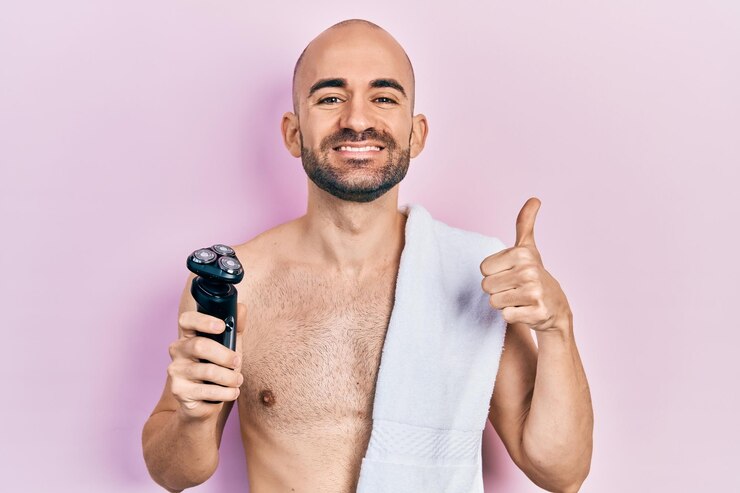 Foto jovem careca sem camisa segurando barbeador elétrico sorrindo feliz e positivo polegar para cima fazendo excelente e sinal de aprovação