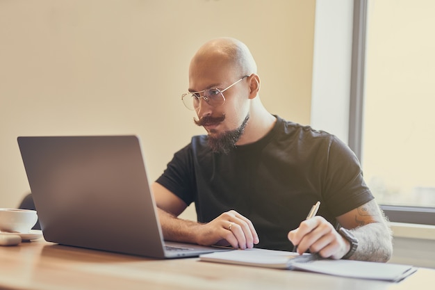 Jovem careca trabalhando em um laptop sentado na mesa, estudando, fazendo algo, anotações, ensino à distância