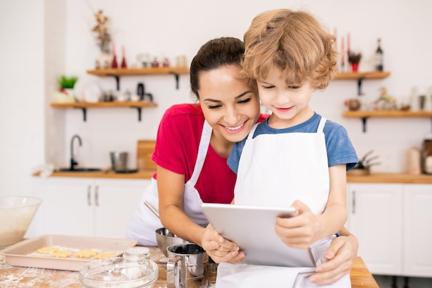 Jovem carinhosa e seu filho com touchpad assistindo receita de vídeo no touchpad enquanto vão preparar o jantar