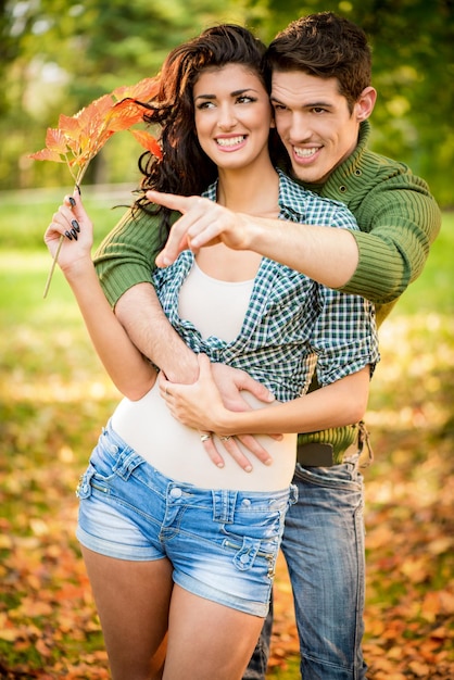 Jovem casal abraçado no parque estendeu os dedos da mão apontando para algo, enquanto a garota carrega na mão um raminho de folhas de outono.