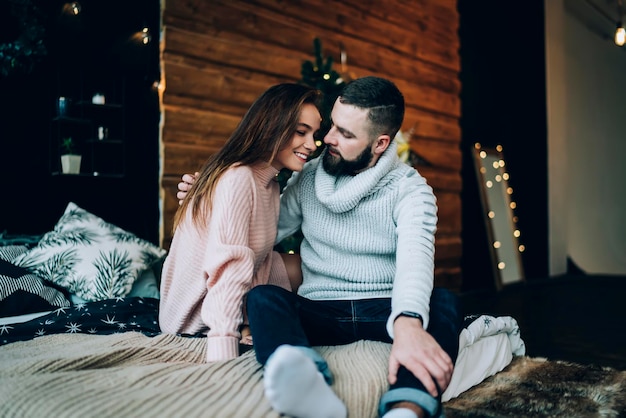 Jovem casal abraçando sentado na cama com decorações de Natal no fundo