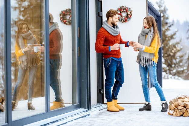 Jovem casal adorável vestido com suéteres coloridos, apreciando a bela vista em pé com bebidas quentes no terraço de uma casa moderna nas montanhas