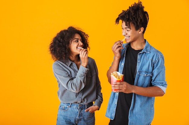 Jovem casal africano alegre parado, isolado, comendo batatas fritas