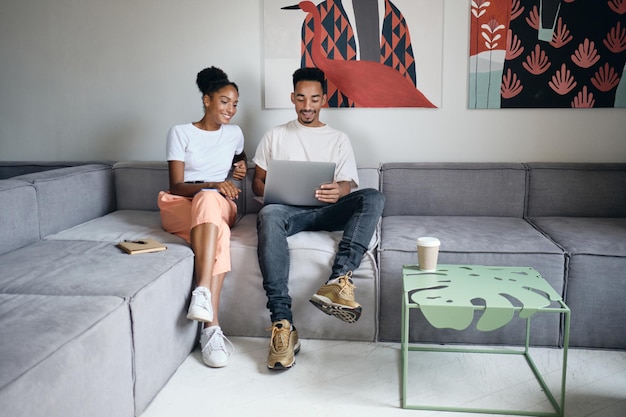 Jovem casal afro-americano casual positivo trabalhando alegremente em laptop juntos no sofá em casa moderna