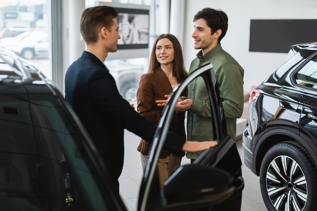 Jovem casal alegre consultando com vendedor de automóveis escolhendo carro novo na concessionária de automóveis