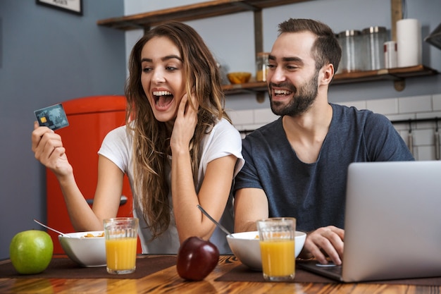 Jovem casal alegre fazendo compras com laptop e cartão de crédito enquanto toma o café da manhã