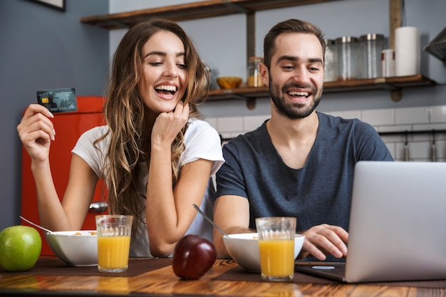 Jovem casal alegre fazendo compras com laptop e cartão de crédito enquanto toma o café da manhã
