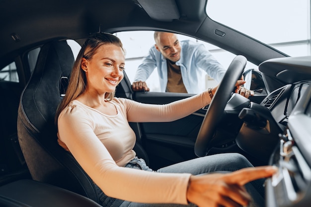 Jovem casal alegre olhando ao redor dentro de um carro novo que vai comprar em uma concessionária de uma loja de automóveis