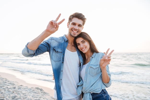 Jovem casal alegre se abraçando e mostrando o símbolo da paz na praia