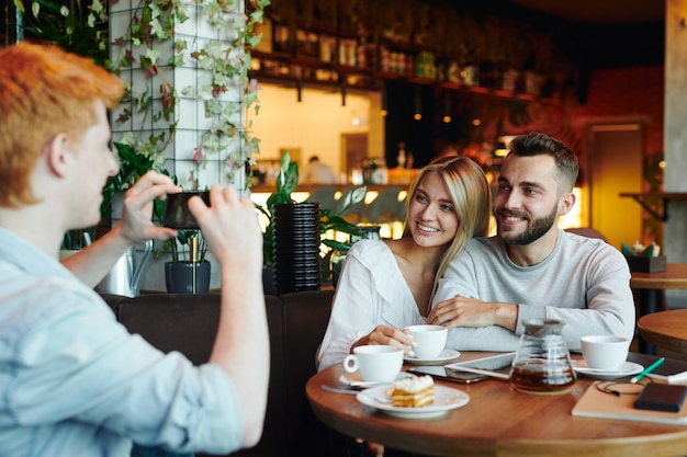 Foto jovem casal alegre sentado à mesa em um café e posando para um amigo de faculdade tirando uma foto deles na câmera do smartphone