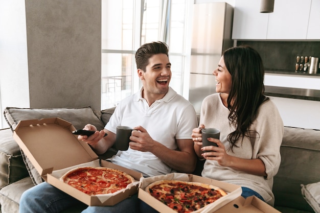Jovem casal alegre sentado em um sofá em casa, comendo pizza