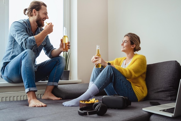 Jovem casal alegre sentado no sofá bebendo cerveja e comendo nachos