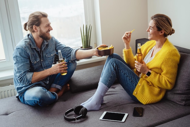 Jovem casal alegre sentado no sofá bebendo cerveja e comendo nachos