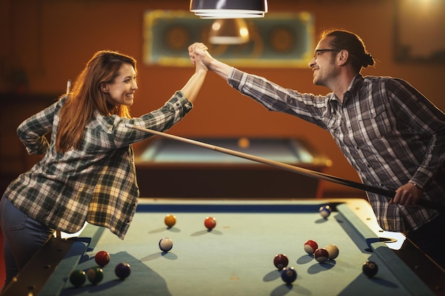 Jovem casal alegre sorridente está jogando bilhar no bar depois do trabalho. Eles estão se divertindo e comemorando a vitória.