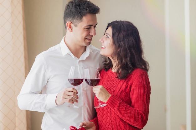 Jovem casal alegre sorrindo um para o outro, bebendo vinho e segurando um pequeno presente branco com fita vermelha nas mãos no restaurante. Conceito de dia dos namorados.
