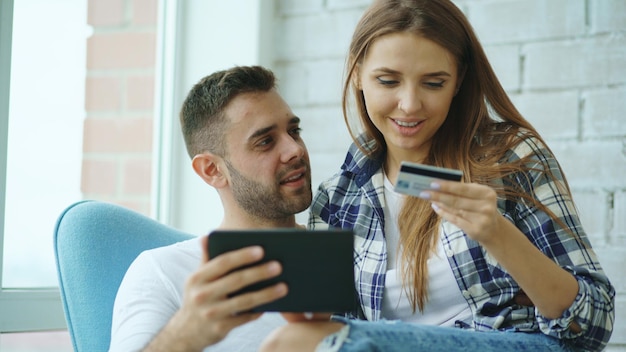 Jovem casal alegre usando computador tablet digital para compras on-line em apartamento moderno