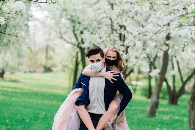 Jovem casal apaixonado andando com máscaras médicas no parque durante a quarentena no dia do casamento.